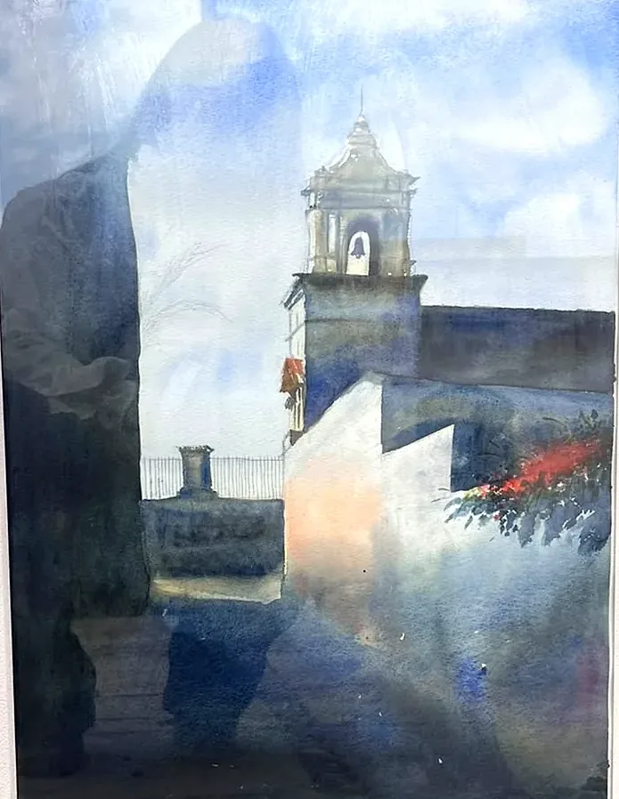 Campanario de sillar visto desde una calle en sombra; el cielo azul lavado ocupa la mitad superior y una mancha de flores rojas rompe el gris del muro de la derecha.